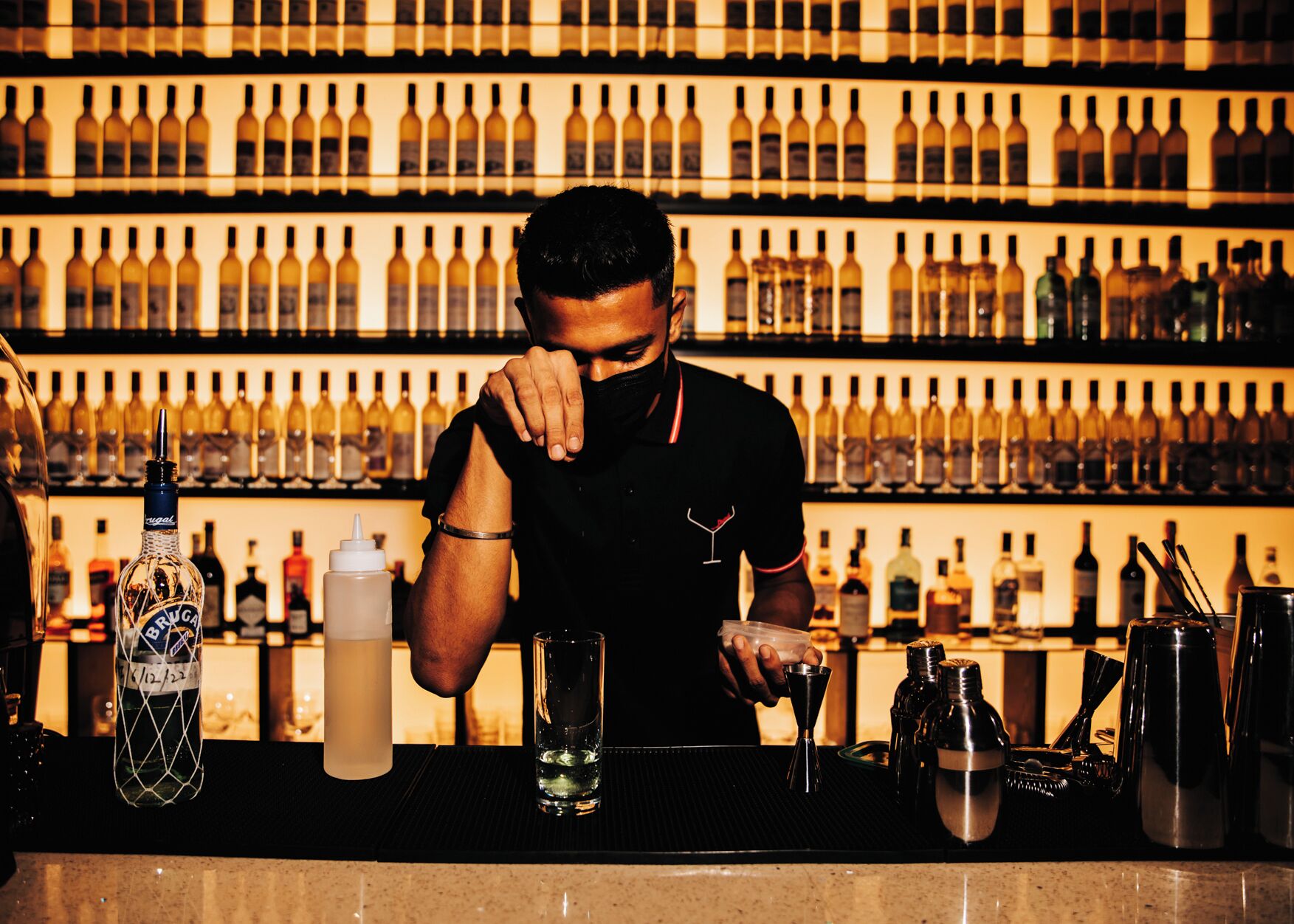 A bartender preparing a drink at a bar with a wall of wine bottles behind him.