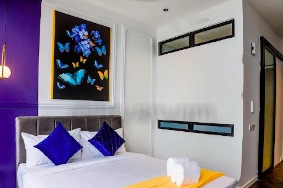 A hotel room with a bed, pillows, a painting of butterflies, a window, and a door.