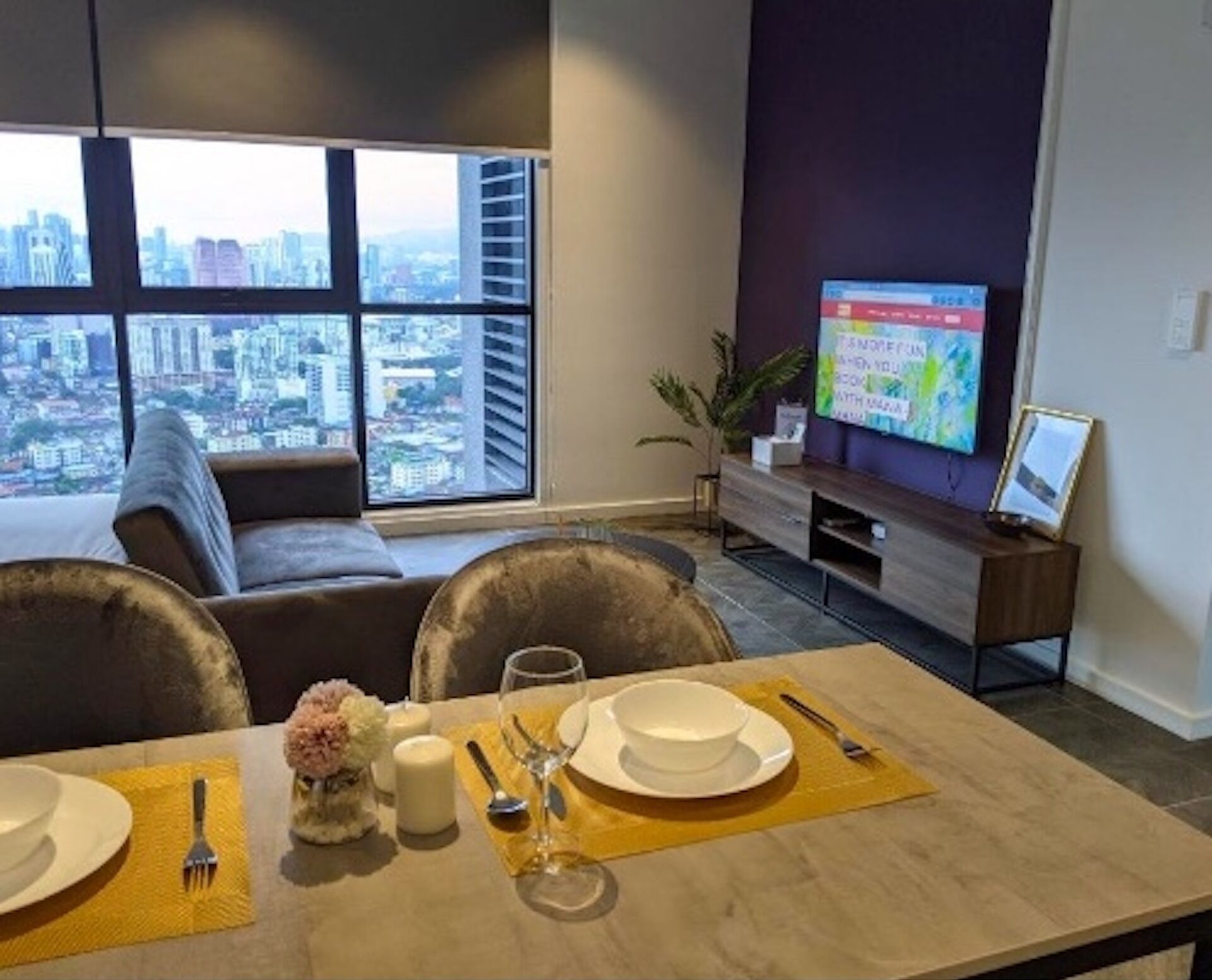 A modern living room with a dining area set for two, a large window with a city view, a flat-screen TV, and a plant.