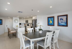 Eine moderne Küche mit weißen Schränken, einem Edelstahlkühlschrank und einem Essbereich mit dunklem Tisch und weißen Stühlen. An der Wand hängen zwei gerahmte Kunstwerke.