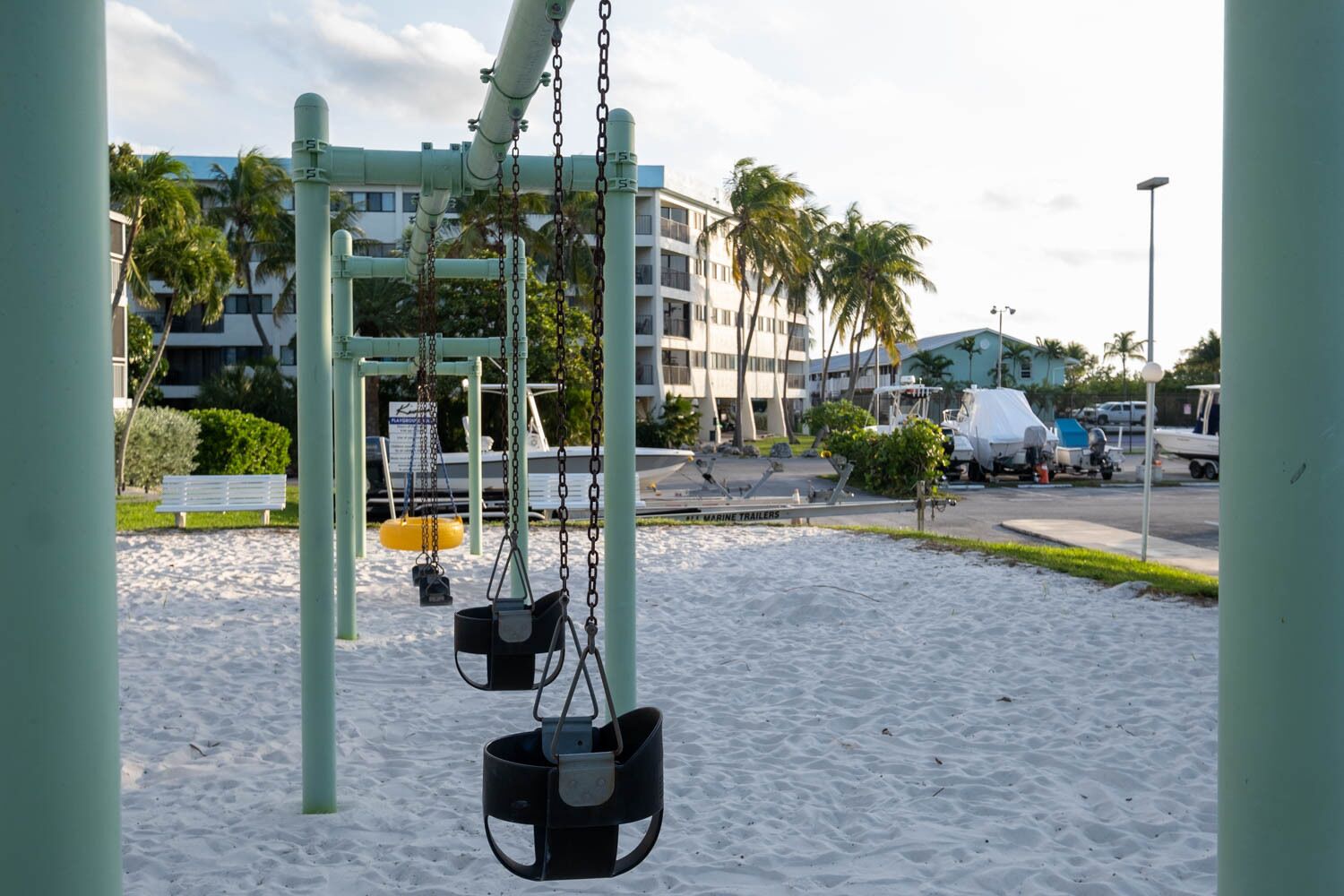 A playground with swings and a sand area, surrounded by buildings and parked boats.