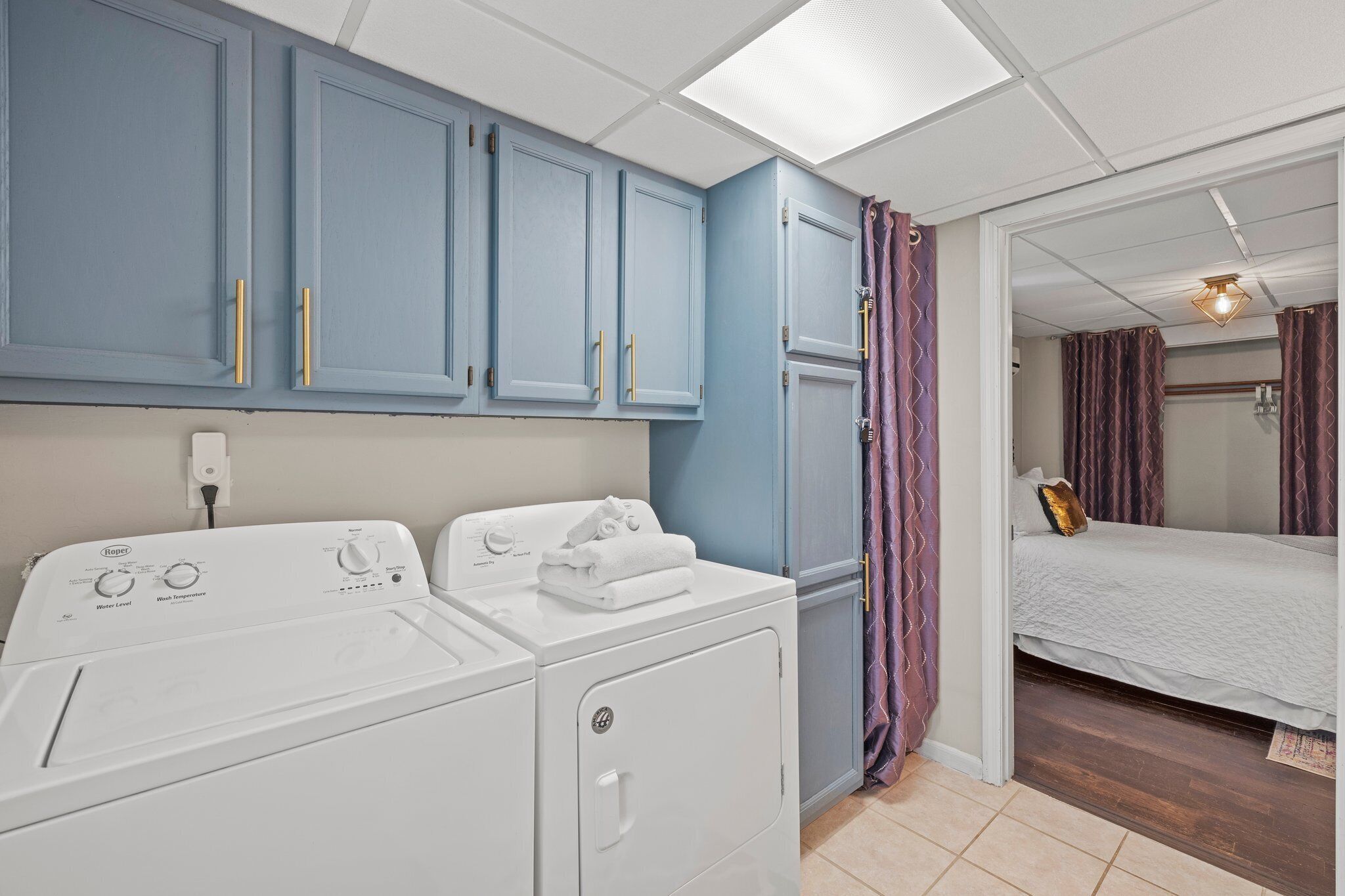 A laundry area with two white washing machines, a blue cabinet with gold handles, and a bed with a white duvet in the adjacent room.