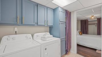 A laundry area with two white washing machines, a blue cabinet with gold handles, and a bed with a white duvet in the adjacent room.