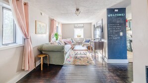 Una sala de estar acogedora con un sofá, una mesita pequeña y una mesa de centro. Hay una pizarra con un mensaje de bienvenida y una lista de servicios.