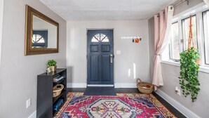 Un pasillo con una puerta azul oscuro, un tapete estampado, un espejo y una planta colgando junto a la ventana.