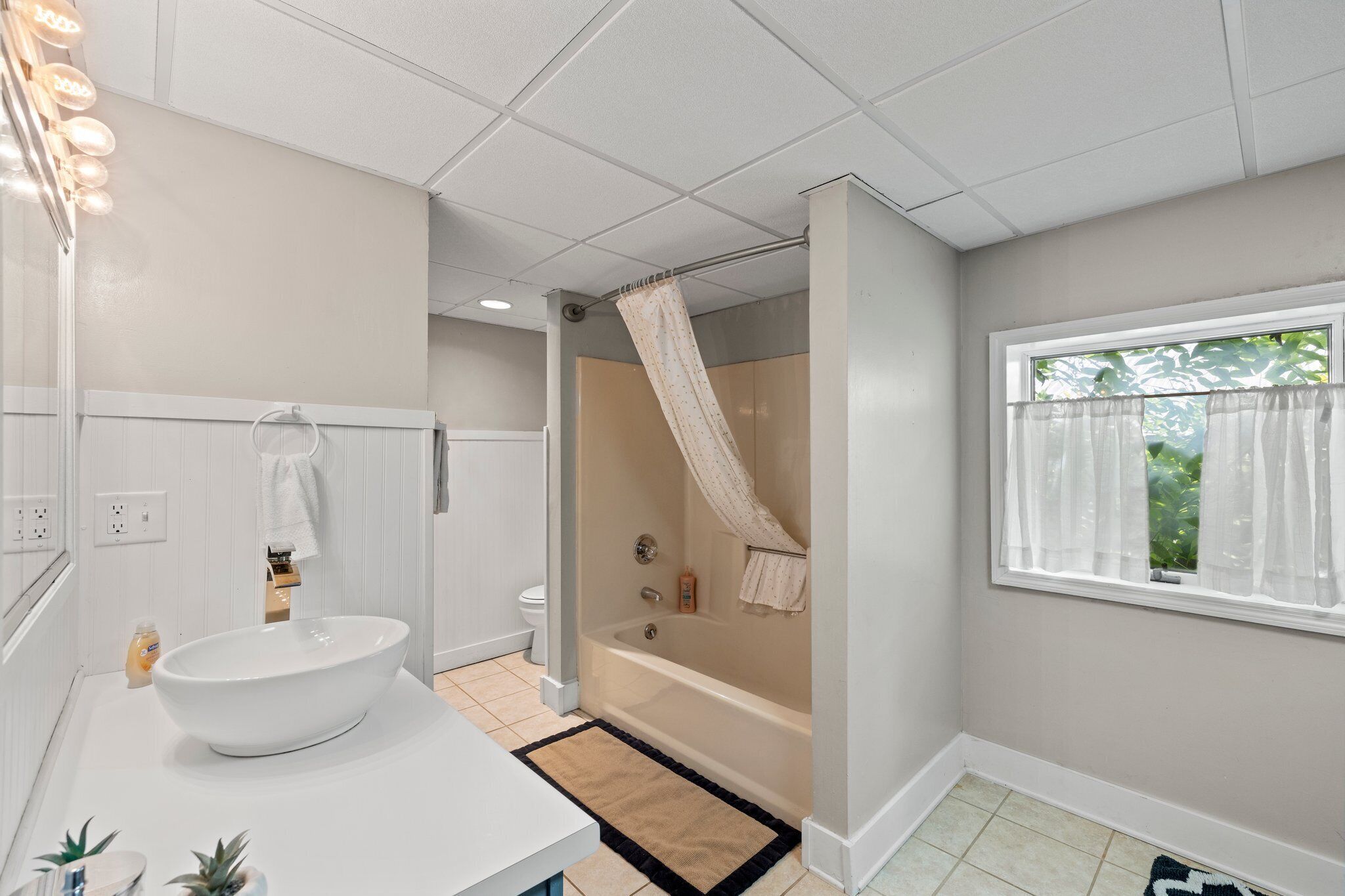 A bathroom with a large white sink, a bathtub, a shower curtain, and a window with curtains.