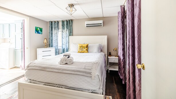 Un dormitorio ordenado con cama, mesitas de noche, un equipo de aire acondicionado y una puerta con cortinas moradas.