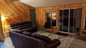 A cozy living room with a leather sofa, a small office chair, a wooden floor, and a sliding glass door leading to a balcony. - Moon Lake Waterfront Getaway (Lewiston)