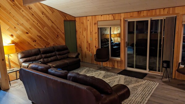 A cozy living room with a leather sofa, a small office chair, a wooden floor, and a sliding glass door leading to a balcony. - Moon Lake Waterfront Getaway (Lewiston)