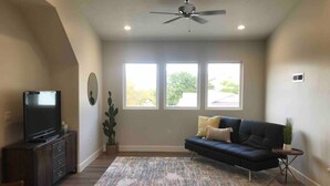 A living room with a dark sofa, a flat-screen TV, a ceiling fan, and a large window with a view of trees. - Private Scenic Stay at Sycamore Lane (Hurricane)
