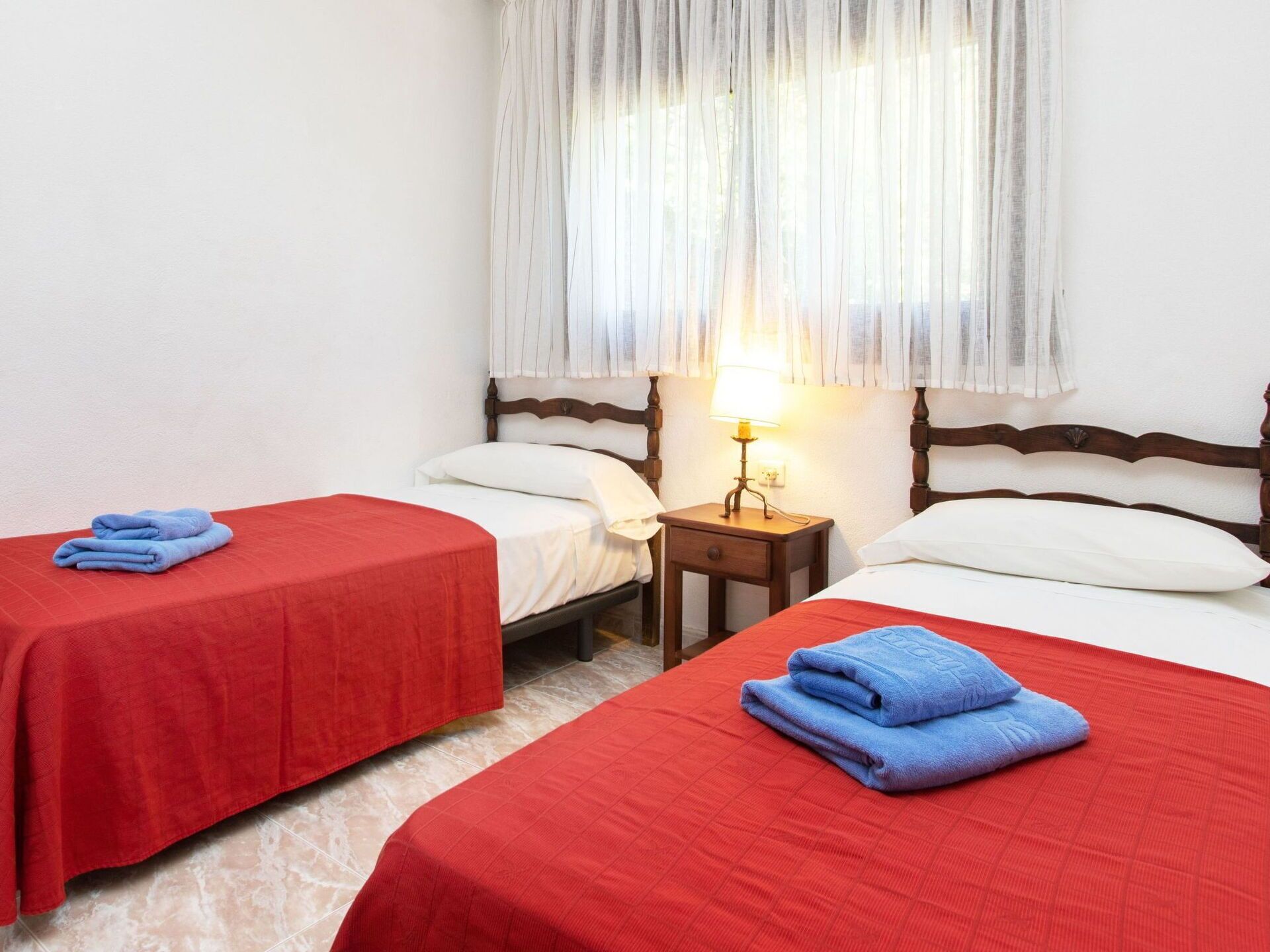 Une chambre d’hôtel avec deux lits simples, chacun recouvert d’une couverture rouge et agrémenté d’une serviette bleue. Une table de chevet en bois avec une lampe se trouve entre les deux lits.