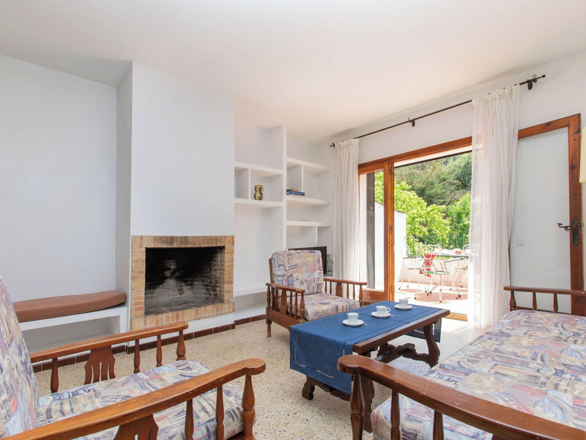 A living room with a fireplace, a dining table with chairs, and a view of the garden through a glass door.
