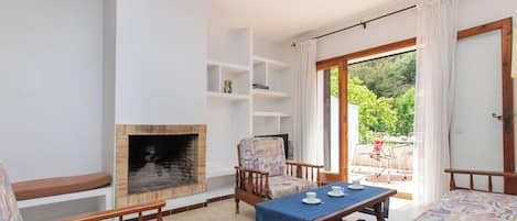 A living room with a fireplace, a dining table with chairs, and a view of the garden through a glass door.