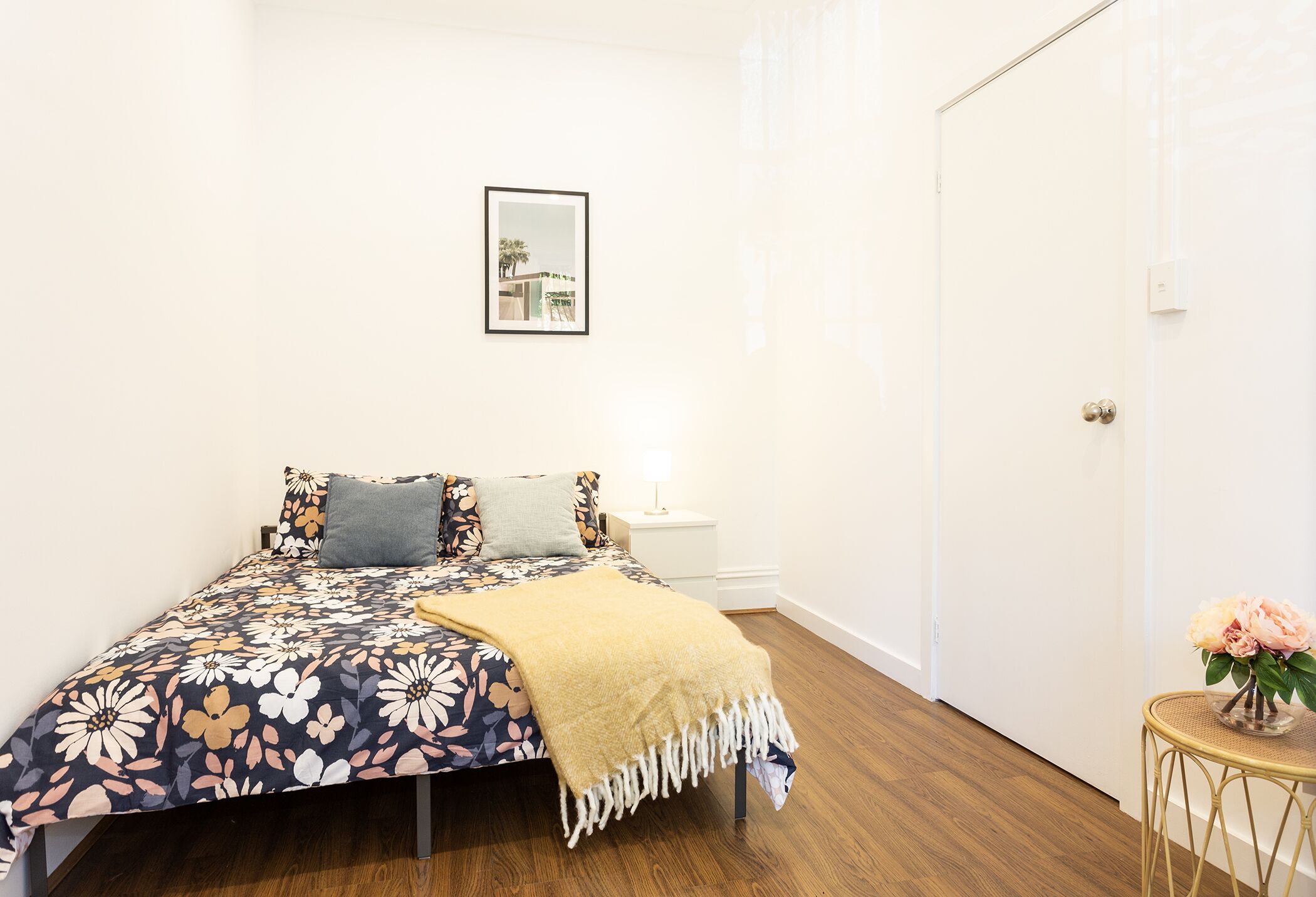 Une chambre à coucher avec une couvre-lit à motifs floraux, une lampe de chevet et un tableau encadré au mur.