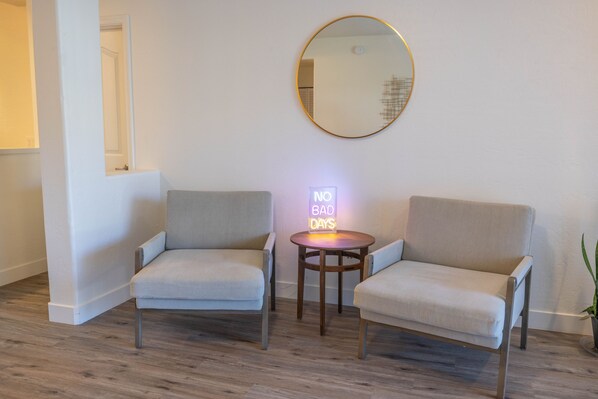 Two light grey armchairs, a round mirror, and a small table with a neon sign.