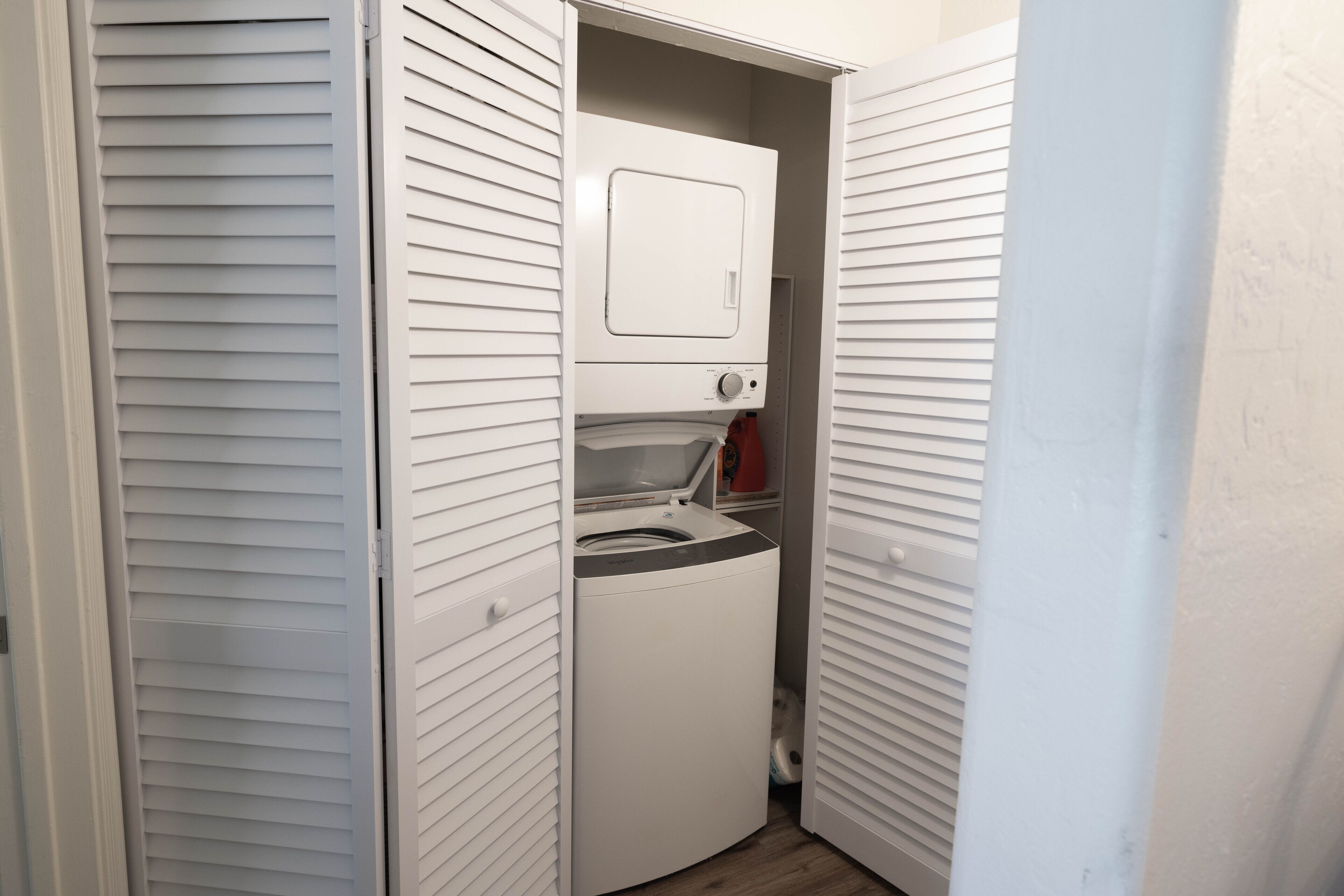 A laundry room with a front-loading washer and dryer, a small shelf with a red container, and a door leading to another room.