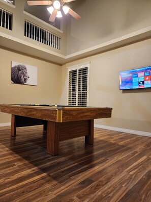 A pool table in a room with wooden flooring, a ceiling fan, a window with blinds, and a television mounted on the wall.