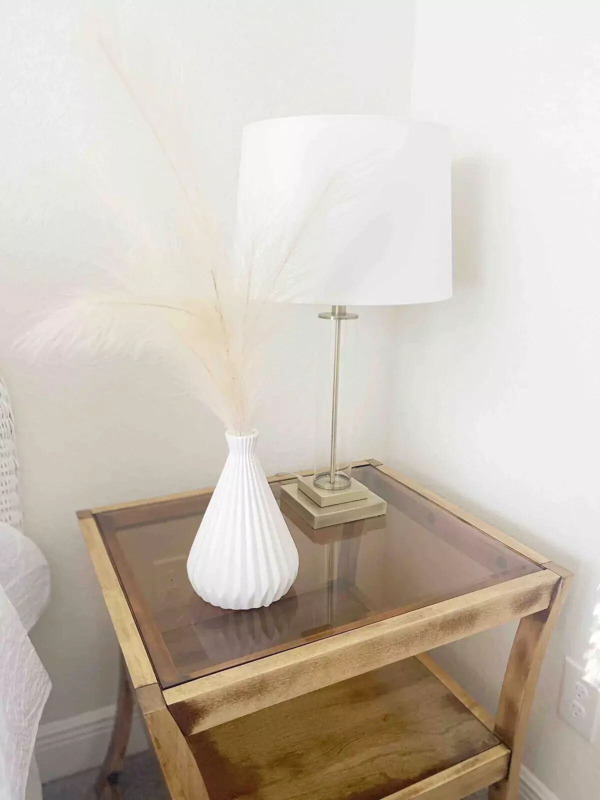 A small wooden side table with a glass top, a white vase with dried flowers, and a floor lamp.