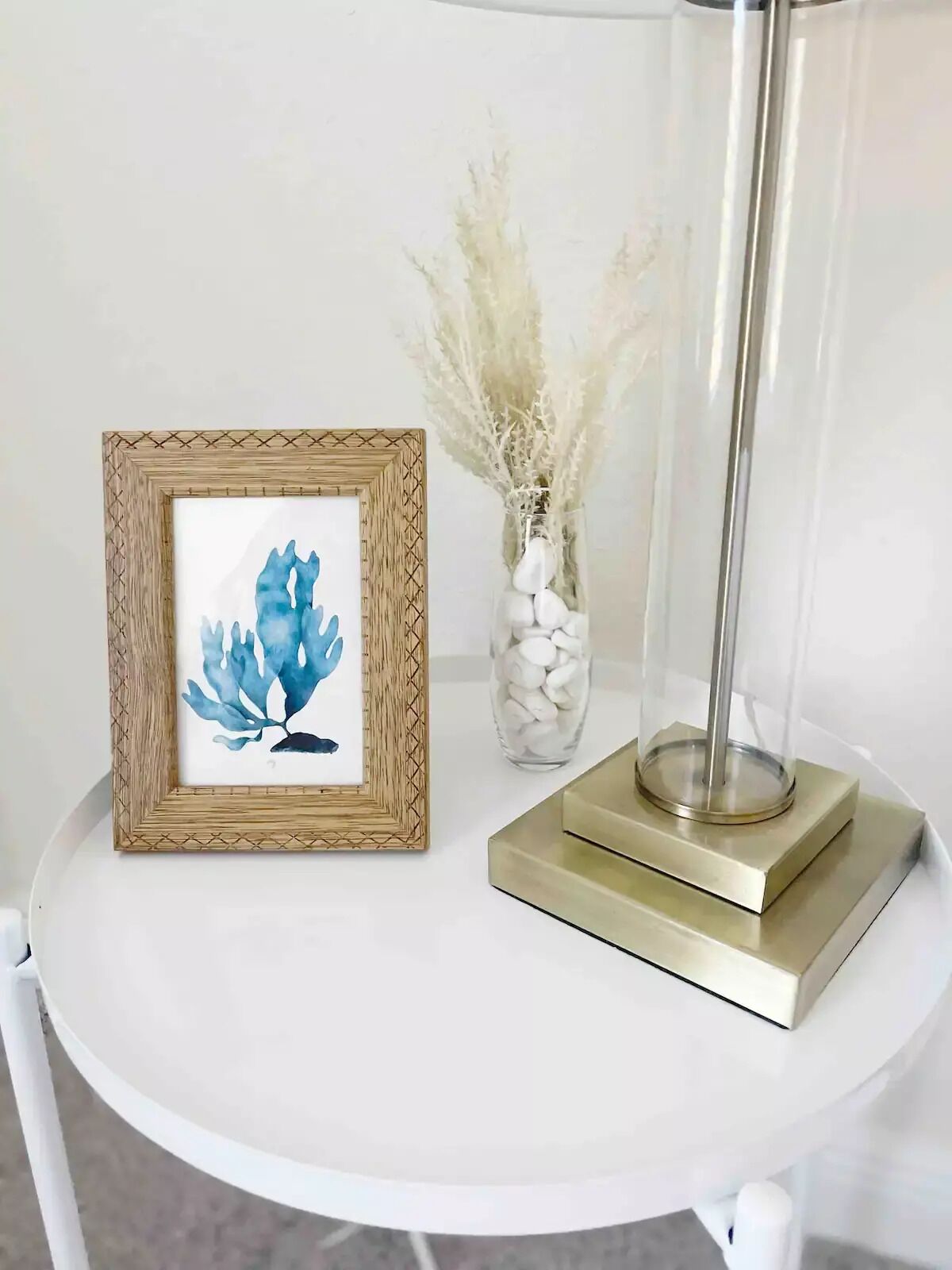 A framed artwork of blue leaves on a white table, a vase with white stones, and a tall floor lamp.