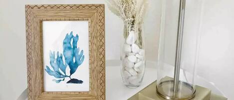 A framed artwork of blue leaves on a white table, a vase with white stones, and a tall floor lamp.