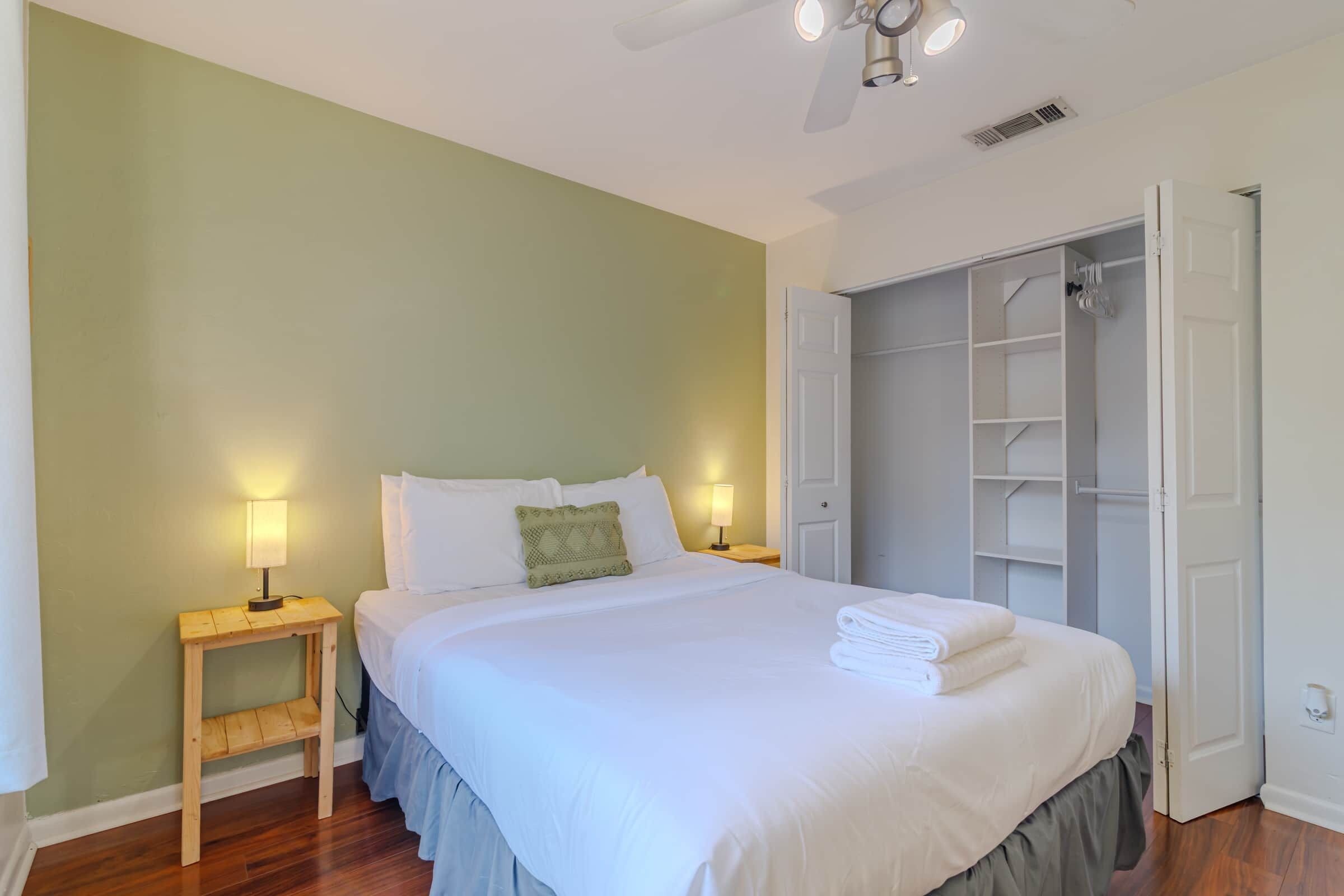 Una cama bien hecha con ropa blanca, una mesita de noche con lámpara y un armario con las puertas abiertas.