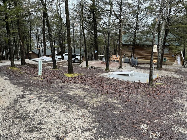 A campground with a cabin labeled "CABIN 3," surrounded by trees and a paved area with a bench and a fire pit.