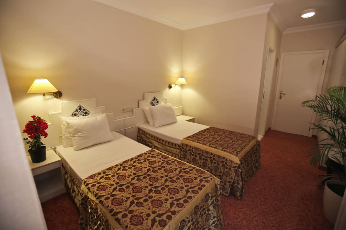 A hotel room with two single beds, a nightstand with a lamp, a potted plant, and a door leading to another room.