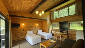 A cabin interior with wooden walls, a sofa, a small table, a TV mounted on the wall, and a window with a view of greenery. - Lakes Lodge Okataina (Lake Okataina)