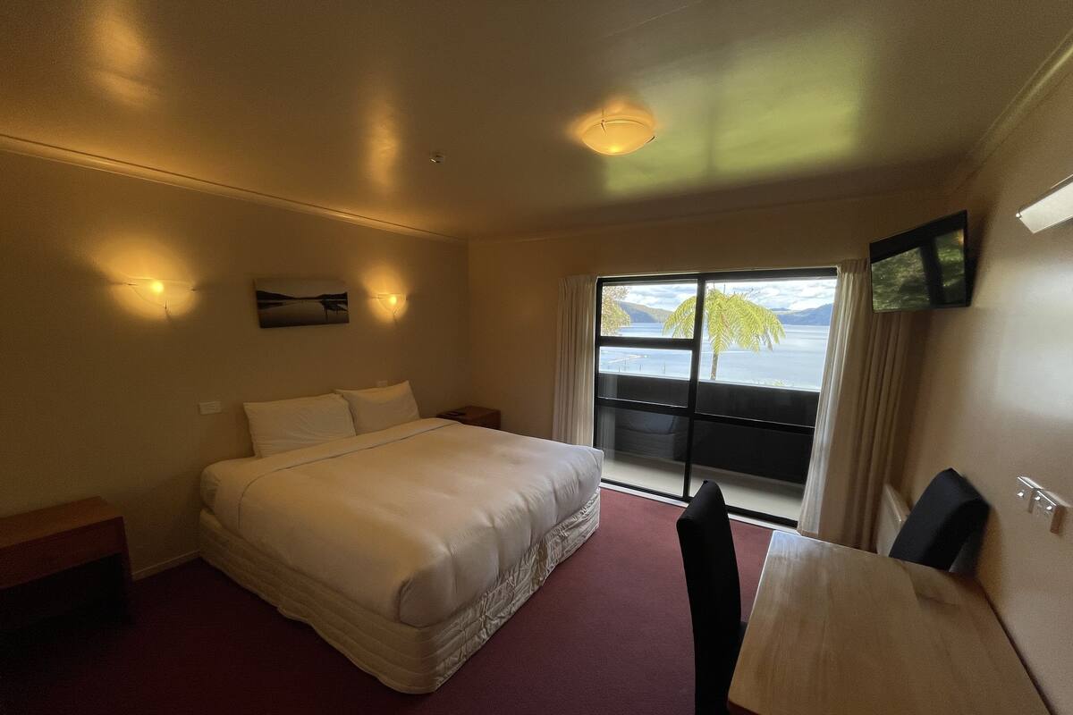 Une chambre d’hôtel comprenant un lit, un bureau, une chaise, une fenêtre donnant sur la mer et un luminaire au plafond.