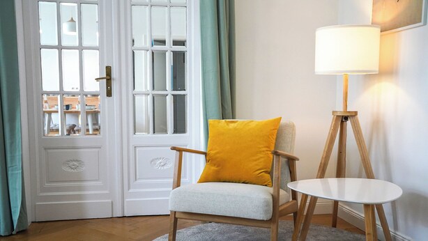 A room with a wooden chair with a yellow cushion, a round white table, a floor lamp, and a white door with glass panels. - Helle und Klare Fewo in Altstadtvilla (Quedlinburg)