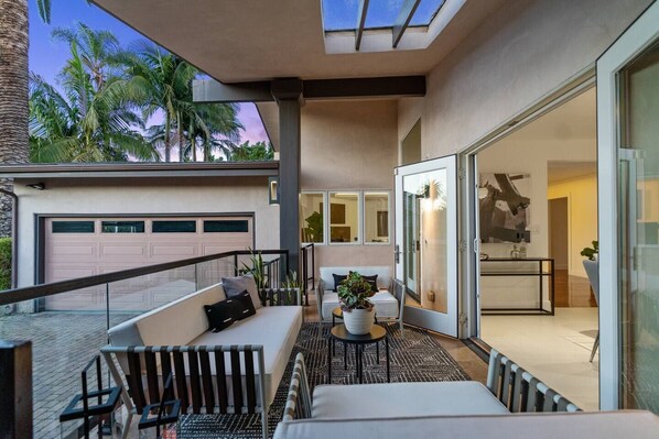 A modern outdoor living area with a sofa, chairs, and a small table, overlooking a garage and a glass door leading to a room.