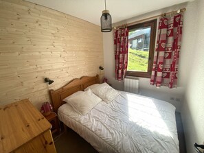 A bedroom with a wooden headboard, a single bed with white bedding, a bedside table, a lamp, and a window with red and white patterned curtains.