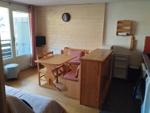 A compact room with a wooden dining table, chairs, a small sofa, a wooden cabinet, and a clock on the wall.