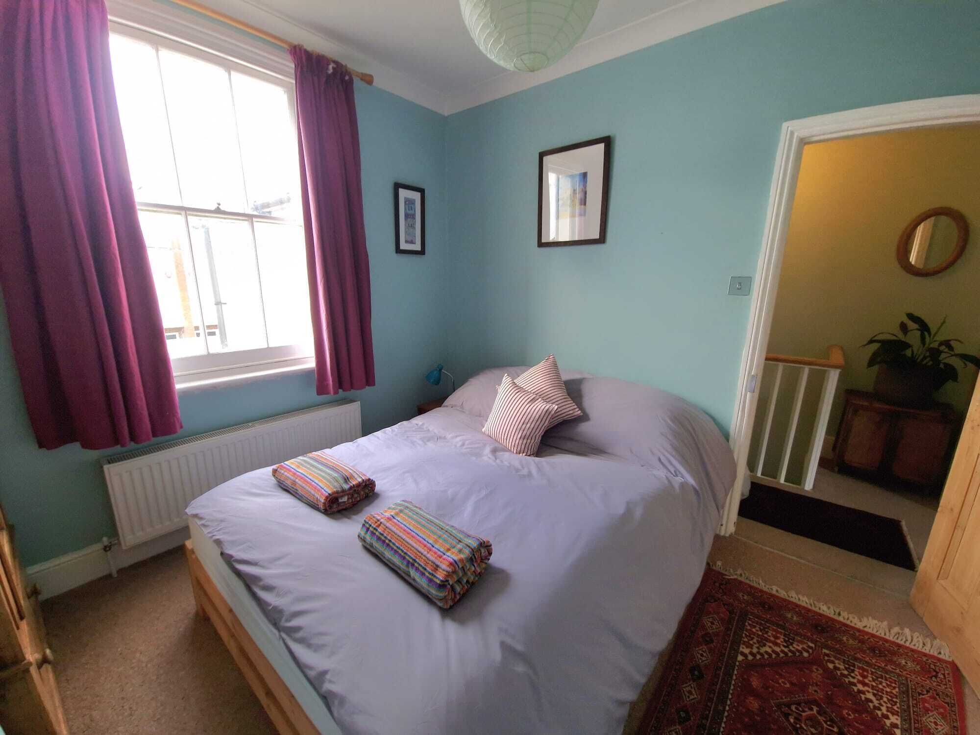 A bedroom with a bed, a window with curtains, a radiator, a framed picture, and a potted plant.