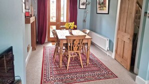 A dining area with a wooden table, chairs, and a patterned rug. There is a window with curtains, a radiator, and framed pictures on the wall. - Delightful 2 bed garden cottage (free parking) (Brighton)