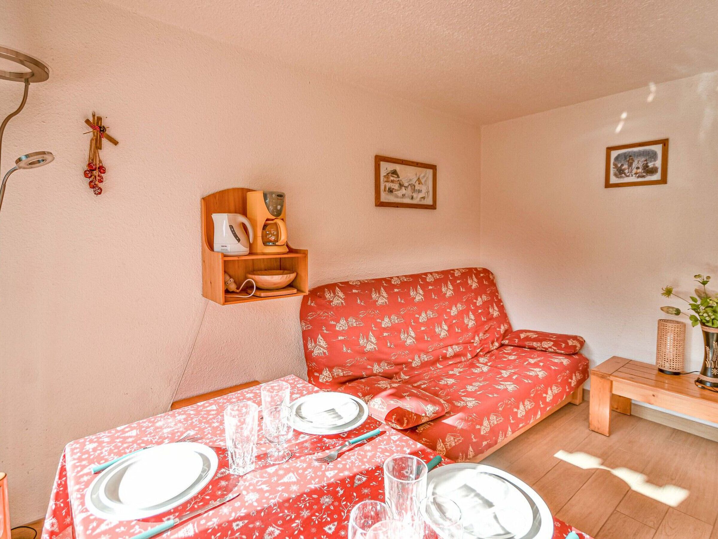 A cozy living room with a red patterned sofa, a wooden shelf with kitchen items, and a dining table set for four.