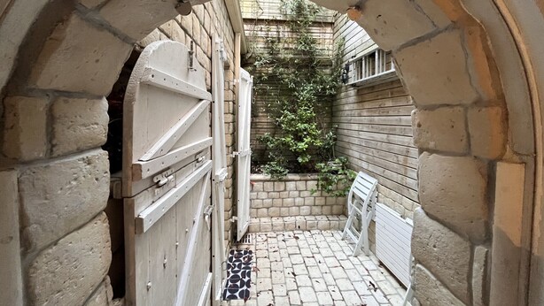 A narrow stone archway leading to a courtyard with a brick floor, a white bench, and a small garden area with climbing plants.