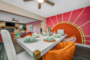 Een eetruimte met een witte tafel gedekt voor zes personen, oranje stoelen en een kleurrijke zonsondergang muurschildering op de muur.