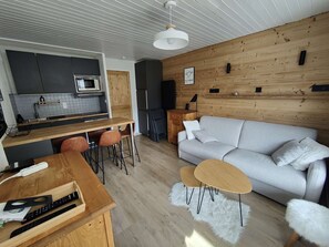 Cozinha moderna com forno embutido e micro-ondas, área de jantar com mesa e cadeiras de madeira, sofá cinza e tapete branco felpudo.