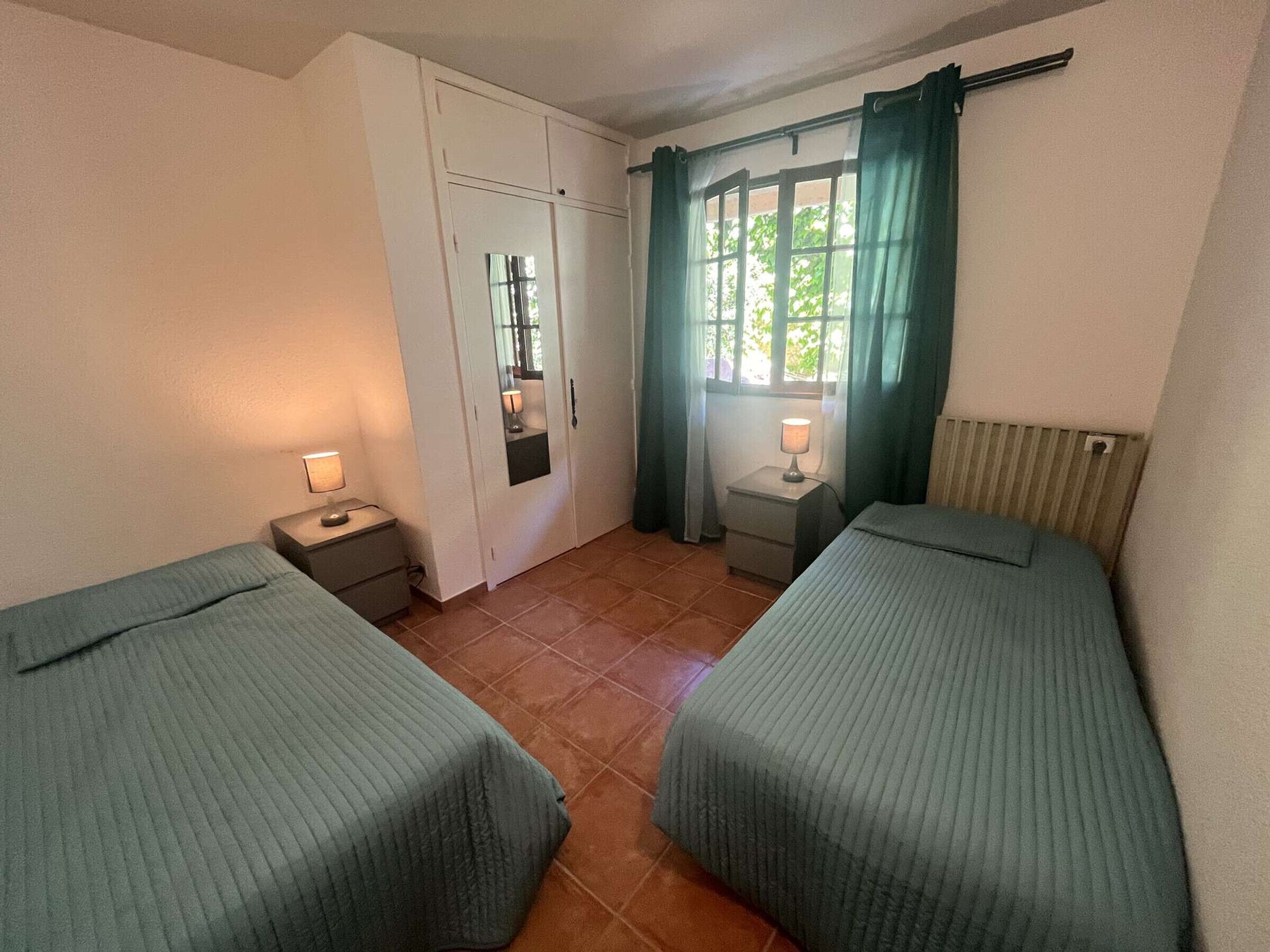 Ein kleines, sauberes Zimmer mit zwei Einzelbetten, einem Kleiderschrank, einem Fenster mit Vorhängen und zwei Nachttischen mit Lampen.