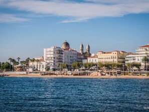 A coastal cityscape with a prominent building featuring a dome and two towers, adjacent to a beach with palm trees and a promenade. - Cozy 2BR Garden Apartment in Saint-Raphaël - Pool, Parking, 600m to Beach (Saint-Raphaël)