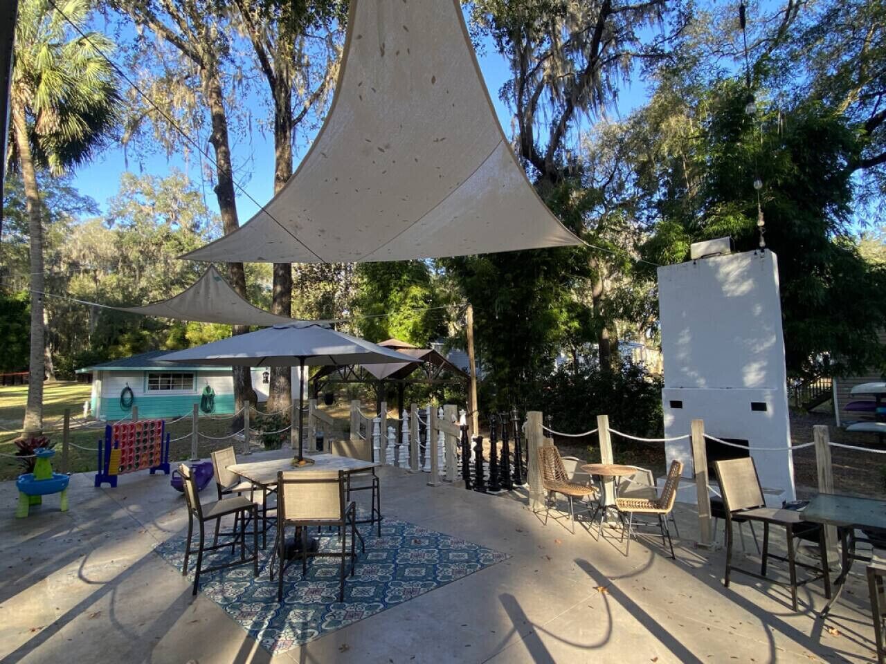 Un espace extérieur aménagé avec des tables et des chaises, protégé par une grande voile d’ombrage et entouré d’arbres.