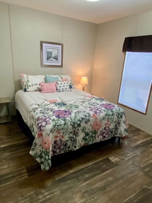 Une chambre à coucher avec une couvre-lit à motifs floraux, une lampe de chevet, un tableau encadré au mur et une fenêtre avec des stores.