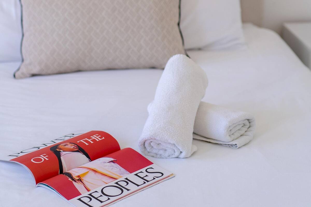 Un lit bien fait, avec des draps blancs, une serviette enroulée et un magazine posé sur le lit.