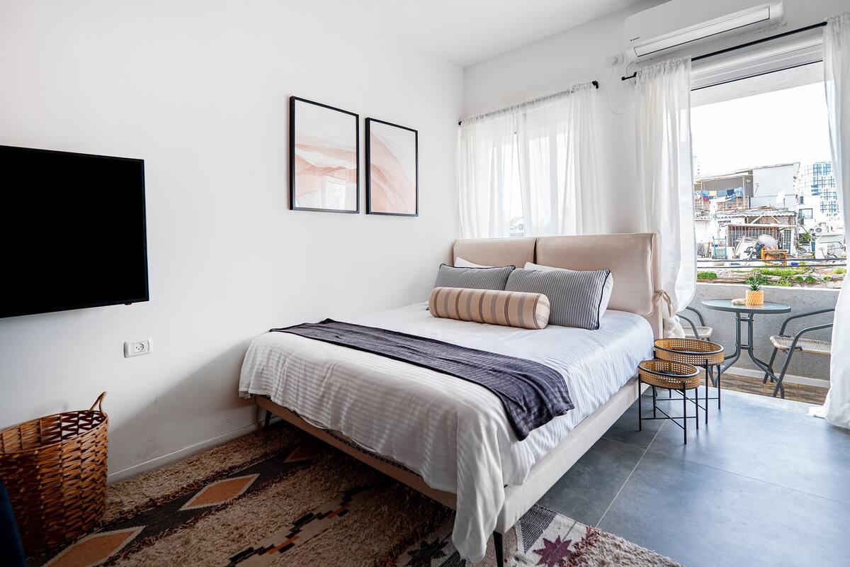 Une chambre avec un lit, un téléviseur à écran plat, une fenêtre avec des rideaux, un petit balcon avec une table et des chaises, et un panier en osier.