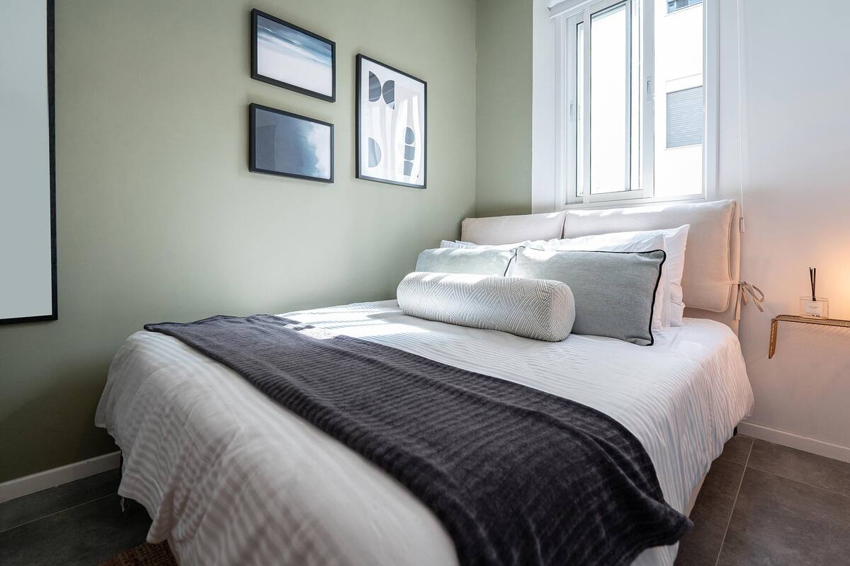 Un lit bien fait, recouvert d’une couette à rayures blanches et grises, d’un plaid gris et agrémenté de plusieurs coussins. Trois œuvres d’art encadrées sont accrochées au mur au-dessus du lit.