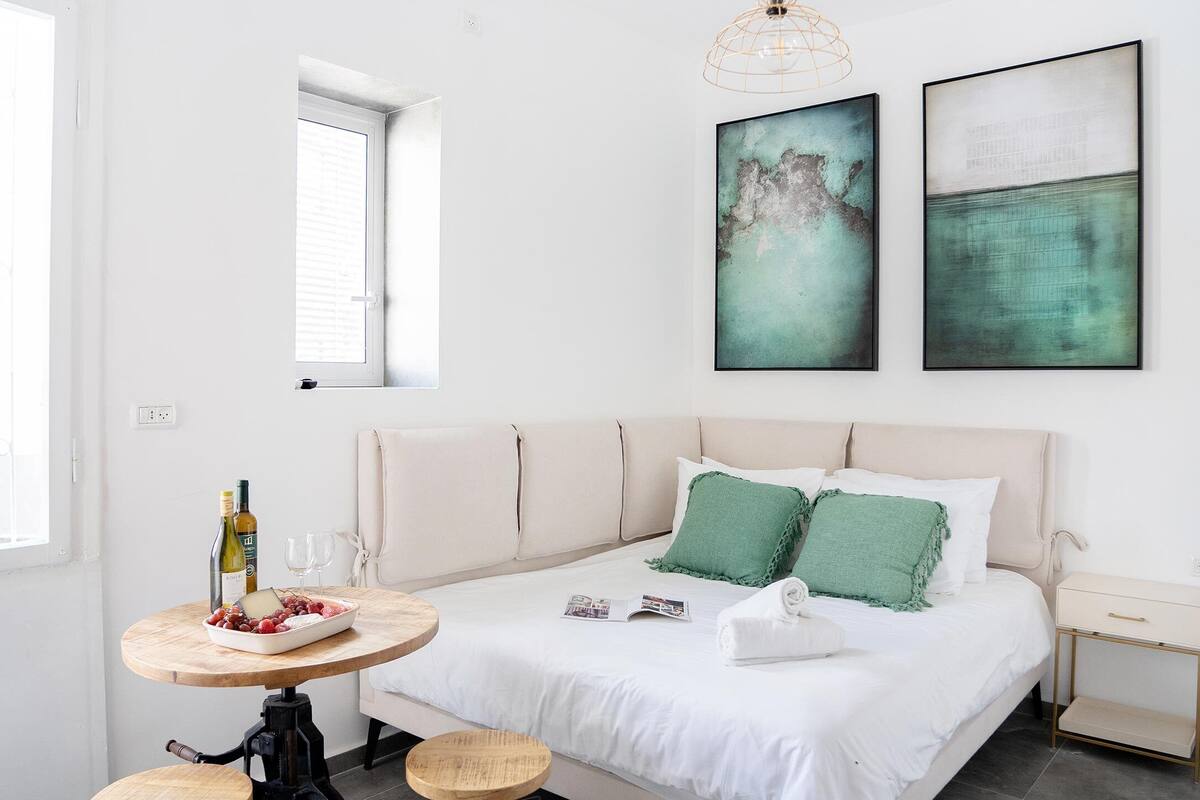 Une chambre à coucher moderne avec un lit, deux tabourets, une table avec des bouteilles et des œuvres d’art accrochées au mur.