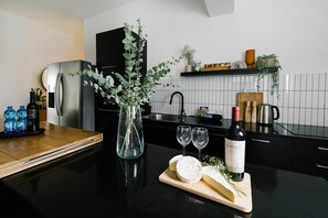 A modern kitchen with a black countertop, a stainless steel refrigerator, a vase with green foliage, a bottle of wine, and a cutting board with cheese. - Carmel Beach House by Berry Stays (Tel Aviv)