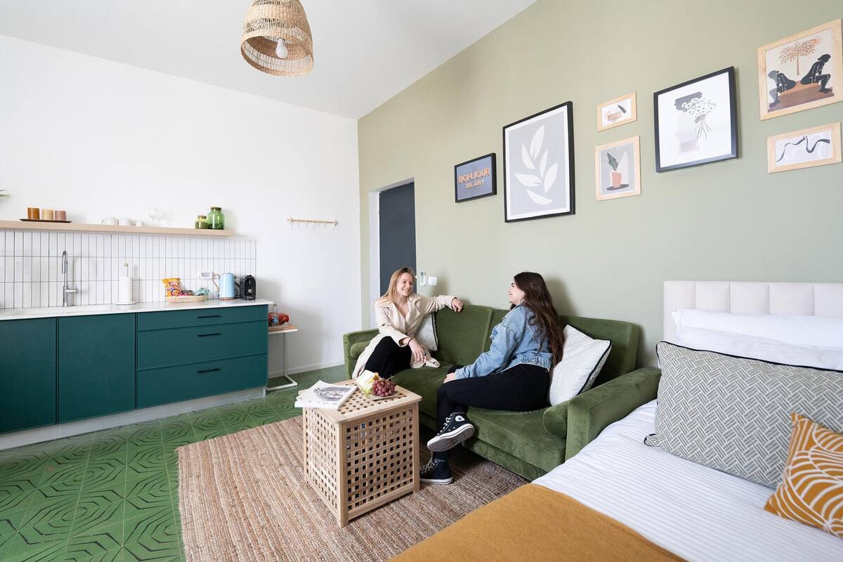 Un salon moderne avec un canapé vert, une petite table d’appoint sur laquelle repose un magazine, et un lit agrémenté d’oreillers à motifs.