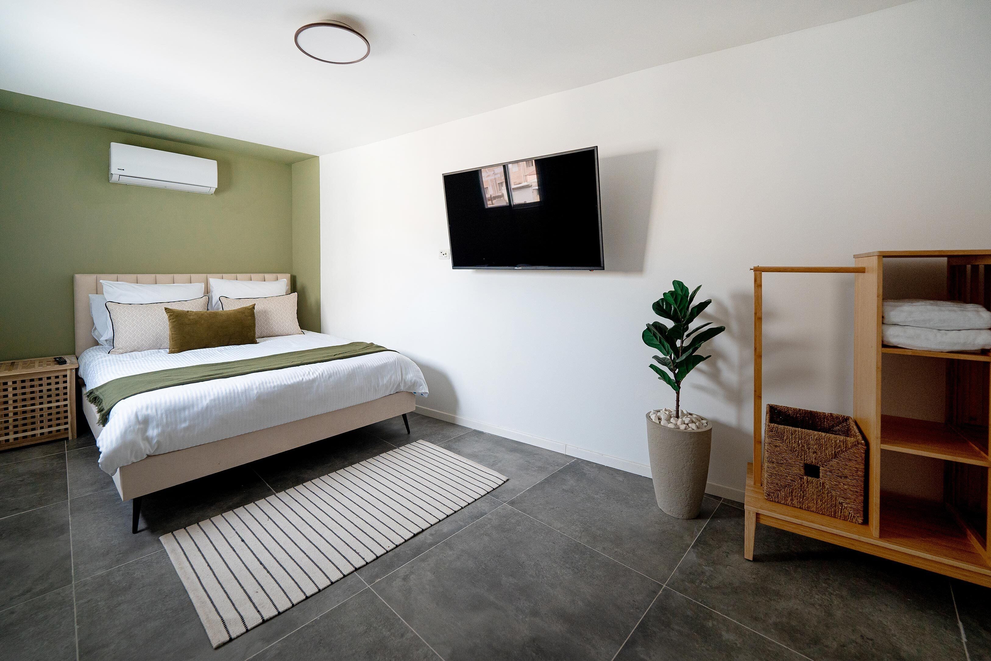 A modern bedroom with a bed, a wall-mounted TV, a potted plant, and a wooden shelf unit.